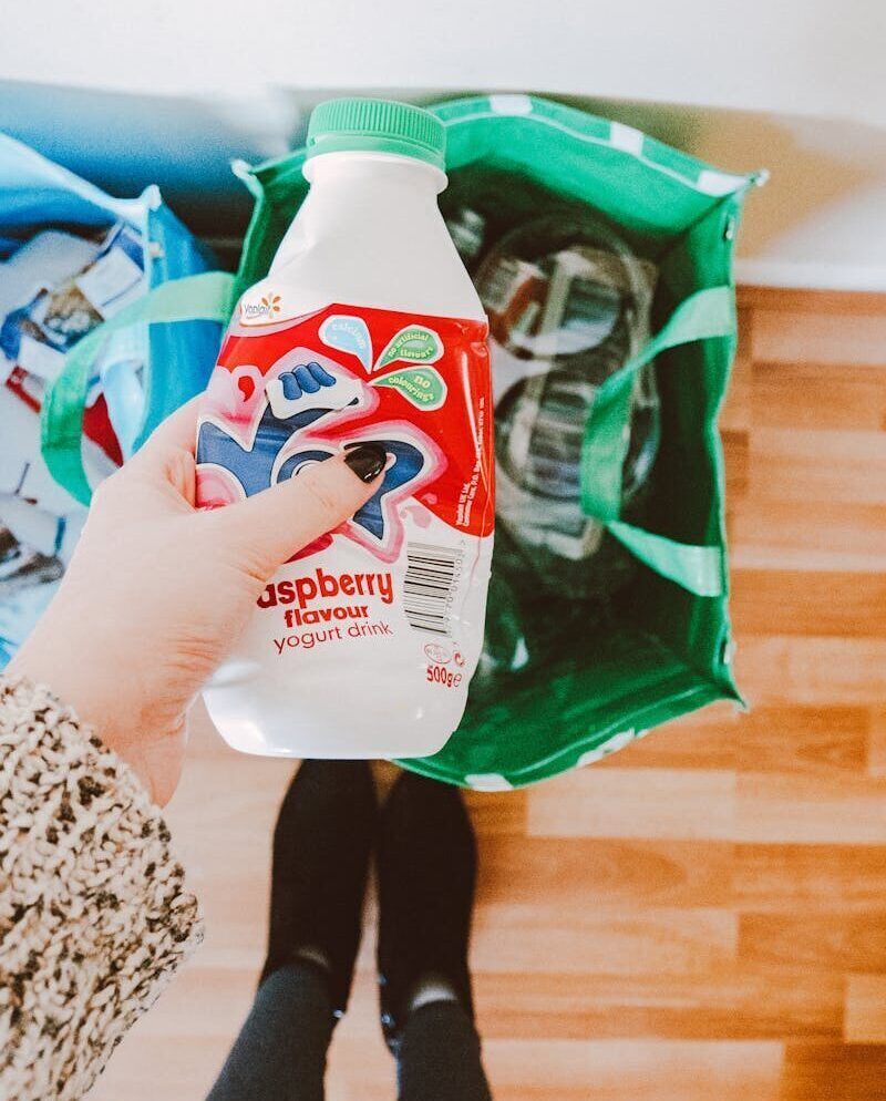 A person recycling a raspberry yogurt drink bottle into a green recycling bag.