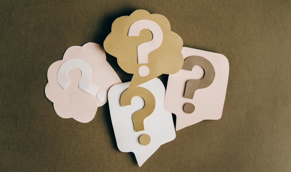 Four paper cutouts of question marks in beige and white on a brown surface, symbolizing inquiry and curiosity.