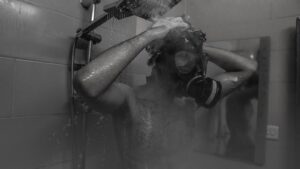 A man wearing a gas mask taking a shower in a monochrome setting.